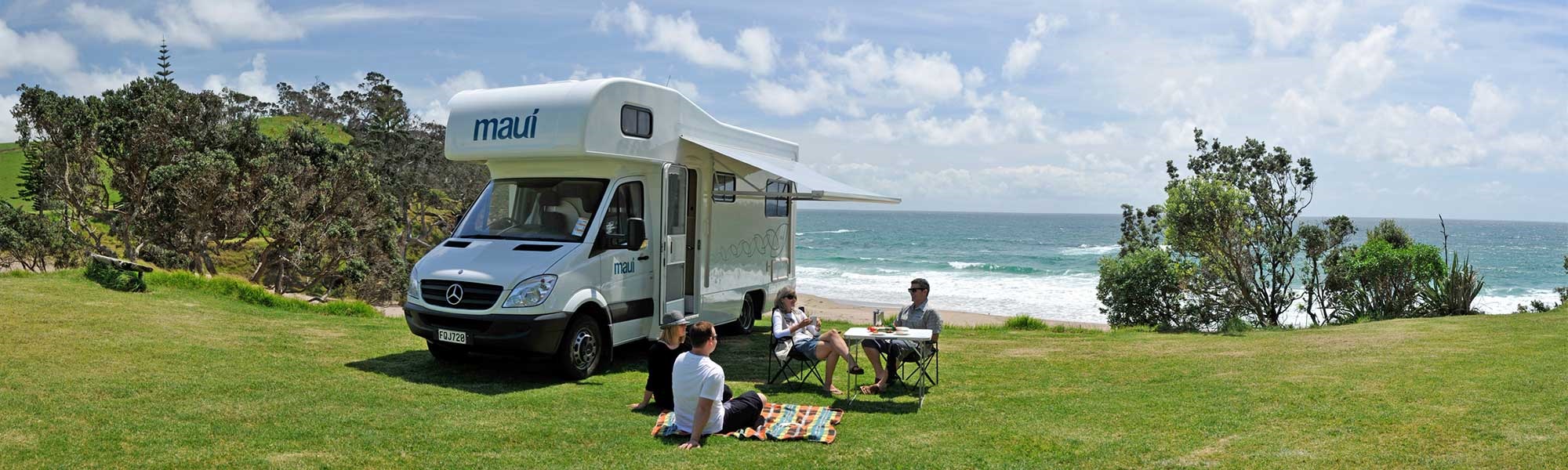 A Maui Campervan in Whananaki, with a family sitting around outside near the beach.