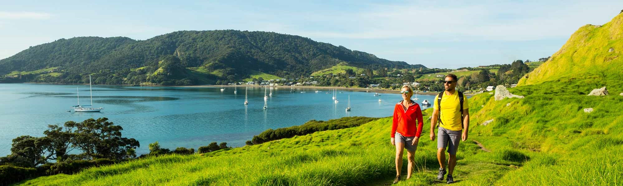 Photo of two people walking on the Smugglers Bay Track with the sea and boats in the background.