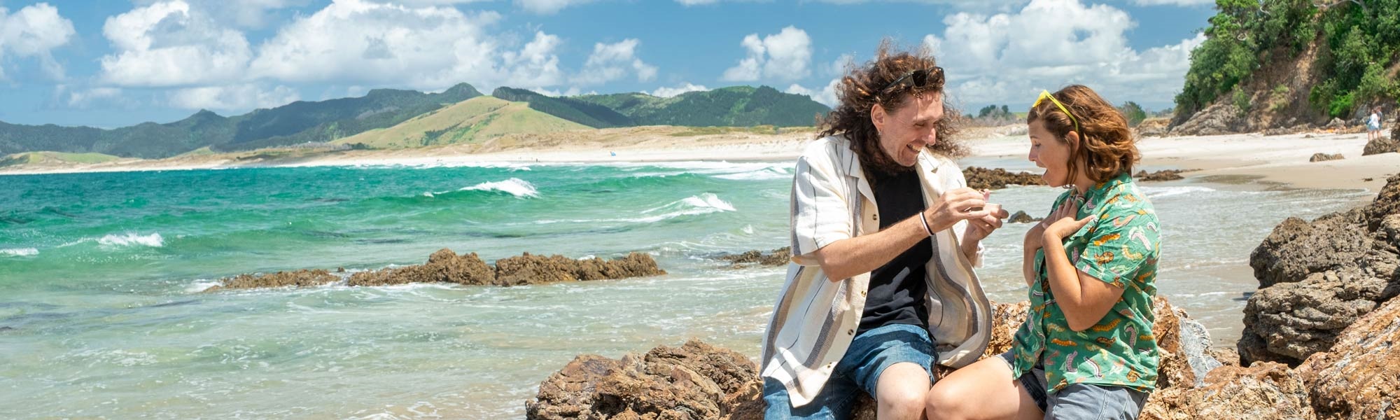 A loving couple proposes marriage on a dramatic seaside landscrape at Kauri Mountain Beach. 