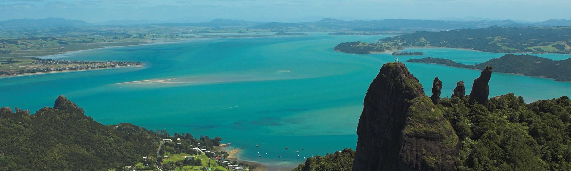 Panoramic view of Whangārei Harbour, with tranquil blue waterways winding between rolling green hills and native bush under a bright, open sky. 