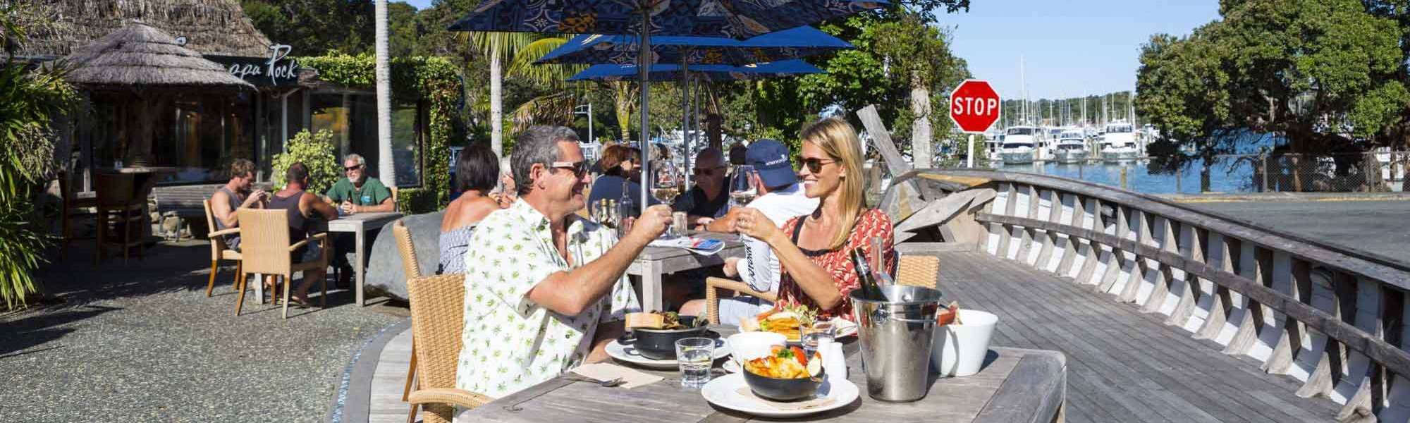 Photo of two people raising their glasses and eating food outside of Schnappa Road at Tutukākā. 